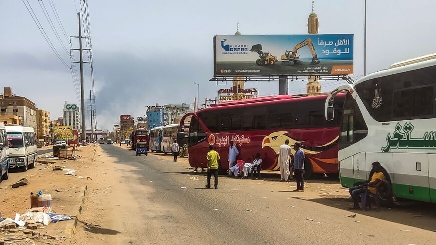 Smoke billows in the distance as people wait next to passenger buses in Sudan's capital Khartoum