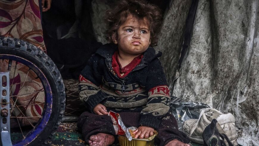 A displaced Palestinian child eats food from a box while sitting in a makeshift tent at a camp beside a street in Rafah