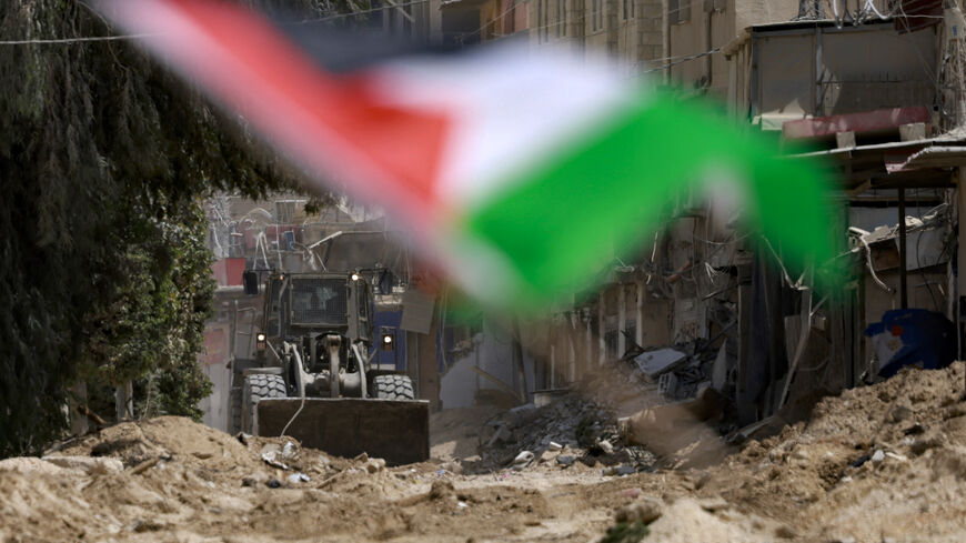 An Israeli army bulldozer digs up a road in the Nur Shams refugee camp in Tulkarem with a Palestinian flag fluttering in the foreground on the second day of a large-scale military operation in the north of the occupied West Bank on August 29, 2024. The death toll climbed on August 29 as the Israeli army said it killed five militants in Tulkarem, bringing to 14 the overall number of people killed since the launch of the West Bank operation the previous day. (Photo by Jaafar ASHTIYEH / AFP) (Photo by JAAFAR A