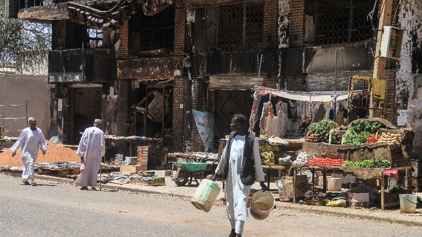 Men walk past street vendors' stands set up beneath a damaged building in the capital Khartoum on the third anniversary of the start of the war 
