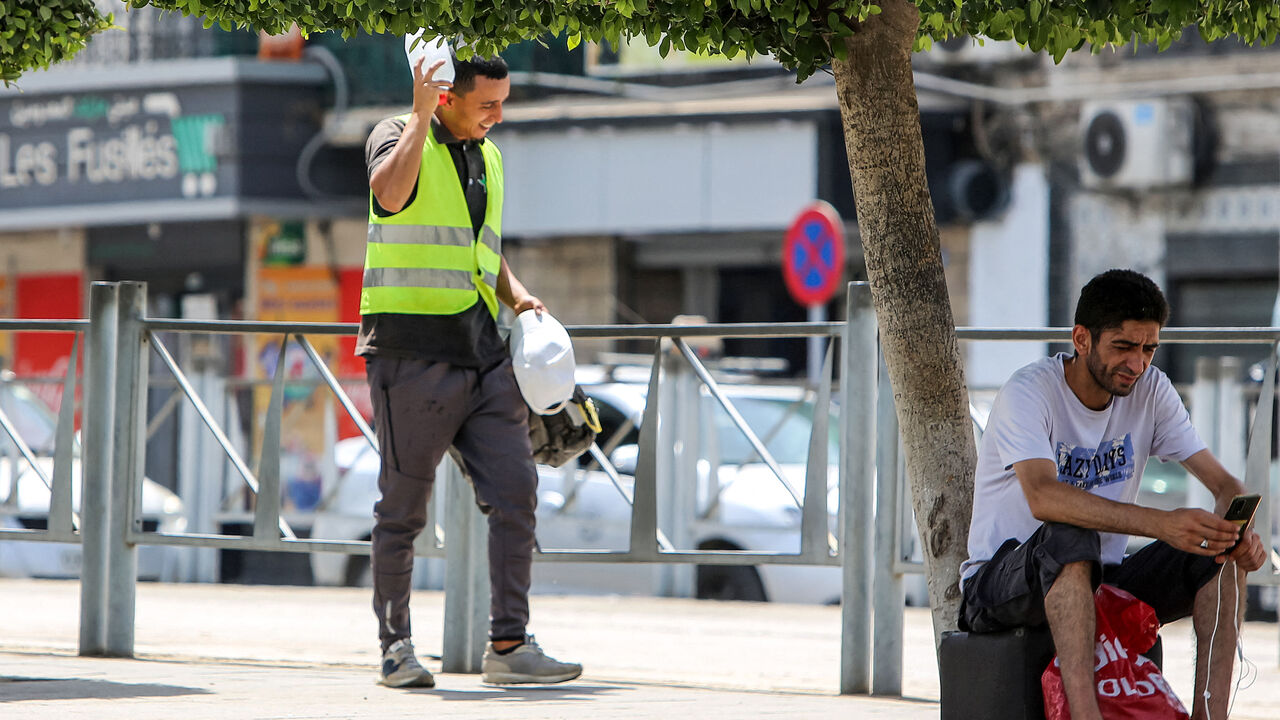 A man browses a phone in the shade of a tree while a construction worker shields his head from the sun with a water bottle while walking along a street in Algiers on July 18, 2023 during a heat wave. (Photo by AFP) (Photo by -/AFP via Getty Images)