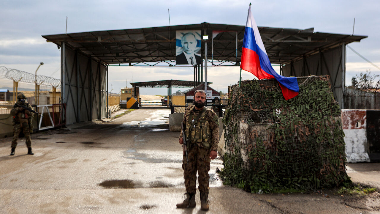 TOPSHOT - A Syrian rebel fighter (C) looks on as he stands at an inspection checkpoint for incoming vehicles before a Russian flag and Russian soldiers behind manning the entrance of the Russian-leased Syrian military base of Hmeimim in Latakia province in western Syria on December 29, 2024. (Photo by AAREF WATAD / AFP) (Photo by AAREF WATAD/AFP via Getty Images)