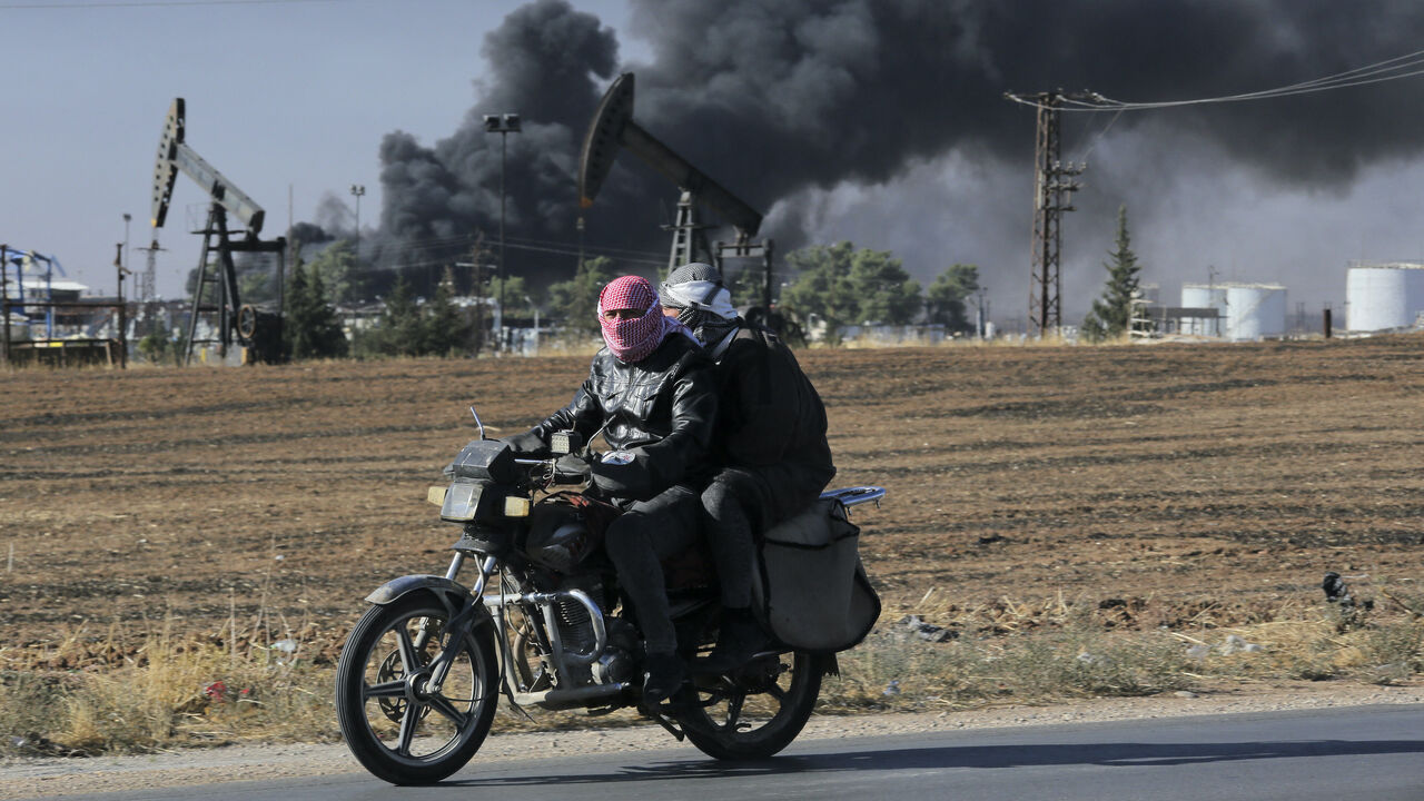 People ride on a motorcycle as thick smoke rises from an oil extracting facility targeted by Turkish shelling near Syria's northeastern border with Turkey in the Qahtaniyah countryside in the far northeast corner of Hasakeh province on Oct. 25, 2024.