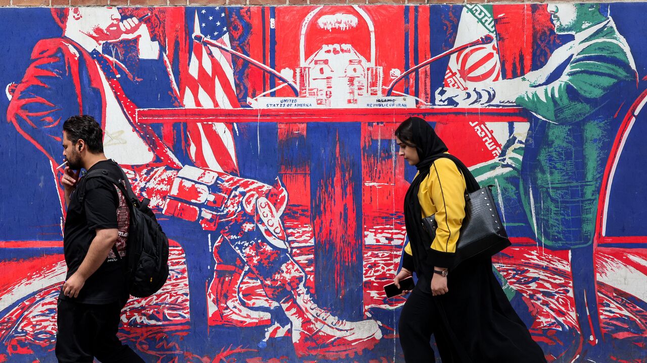 A man and woman walk past a mural depicting an Iranian official in civilian clothing sitting across the table from an American official wearing military fatigues painted on the outer walls of the former US embassy in Tehran, colloquially referred to as the "Spy Den," on April 8, 2025.