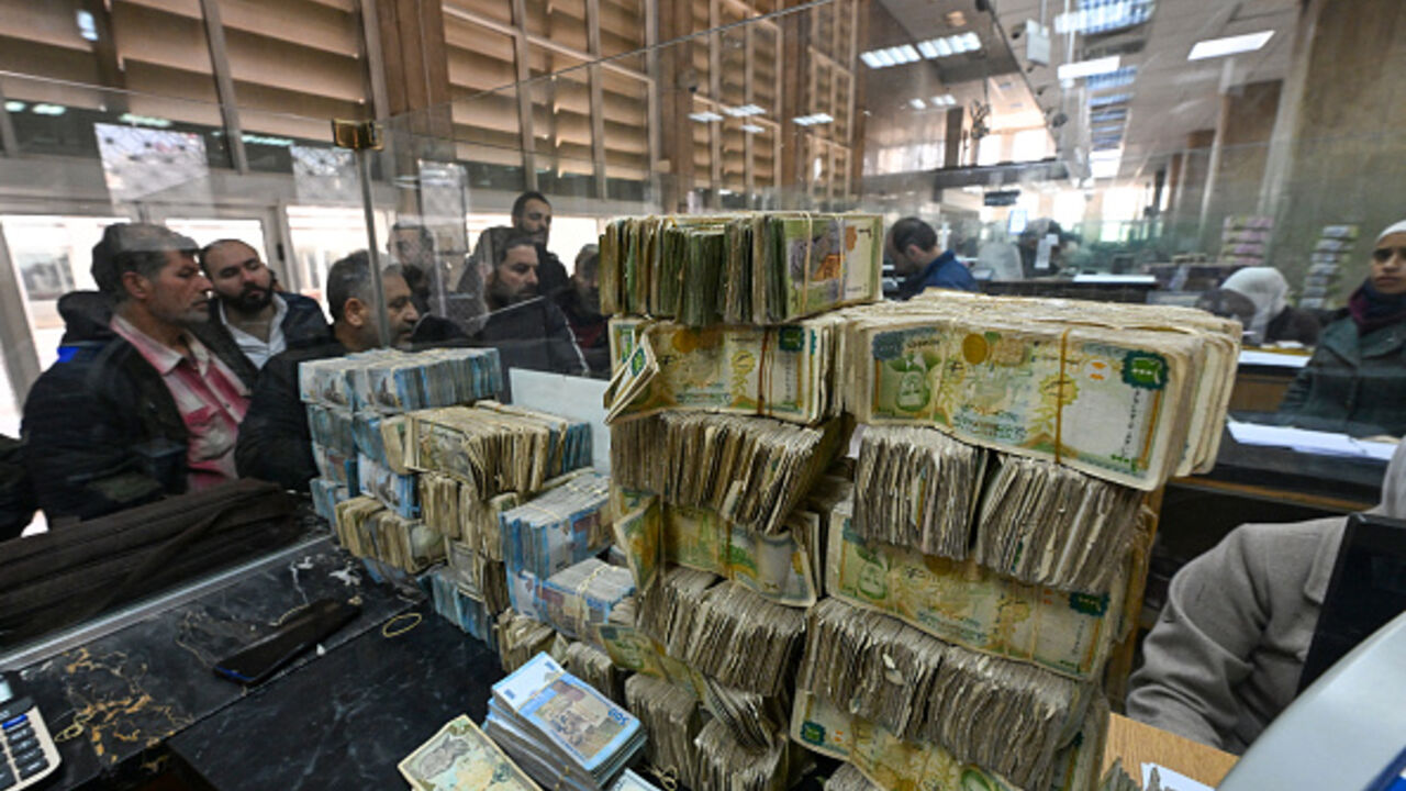 People queue in front of a teller next to a pile of banknotes of different currencies at a commercial bank affiliated with Syria's central bank in Damascus on December 16, 2024. Governments worldwide are stepping up efforts to engage with Syria's new interim rulers, just over a week after Islamist-led rebels ousted president Bashar al-Assad, ending decades of brutal rule and civil war. (Photo by Louai Beshara / AFP) / "The erroneous CAPTION appearing in the metadata of this photo by Louai Beshara has been