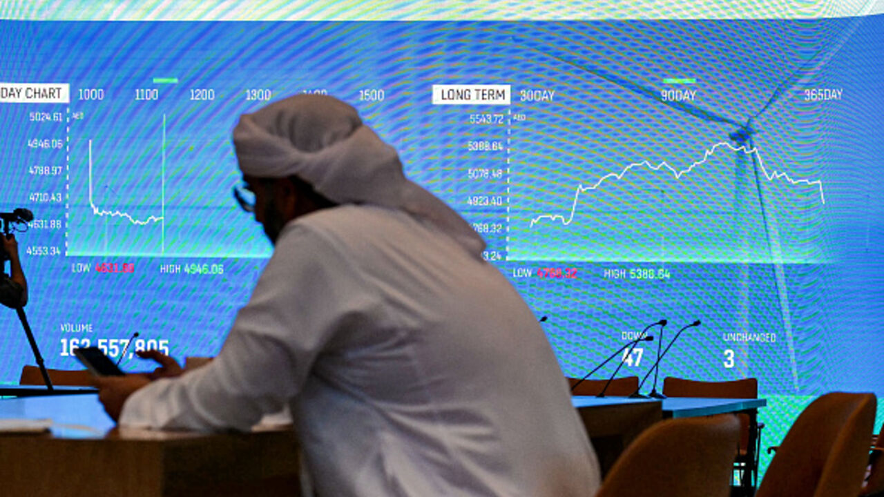 A man browses his phone while sitting near a screen displaying the stock indices at the Dubai Financial Market (DFM) stock exchange in Dubai on April 7, 2025. (Photo by Giuseppe CACACE / AFP) (Photo by GIUSEPPE CACACE/AFP via Getty Images)