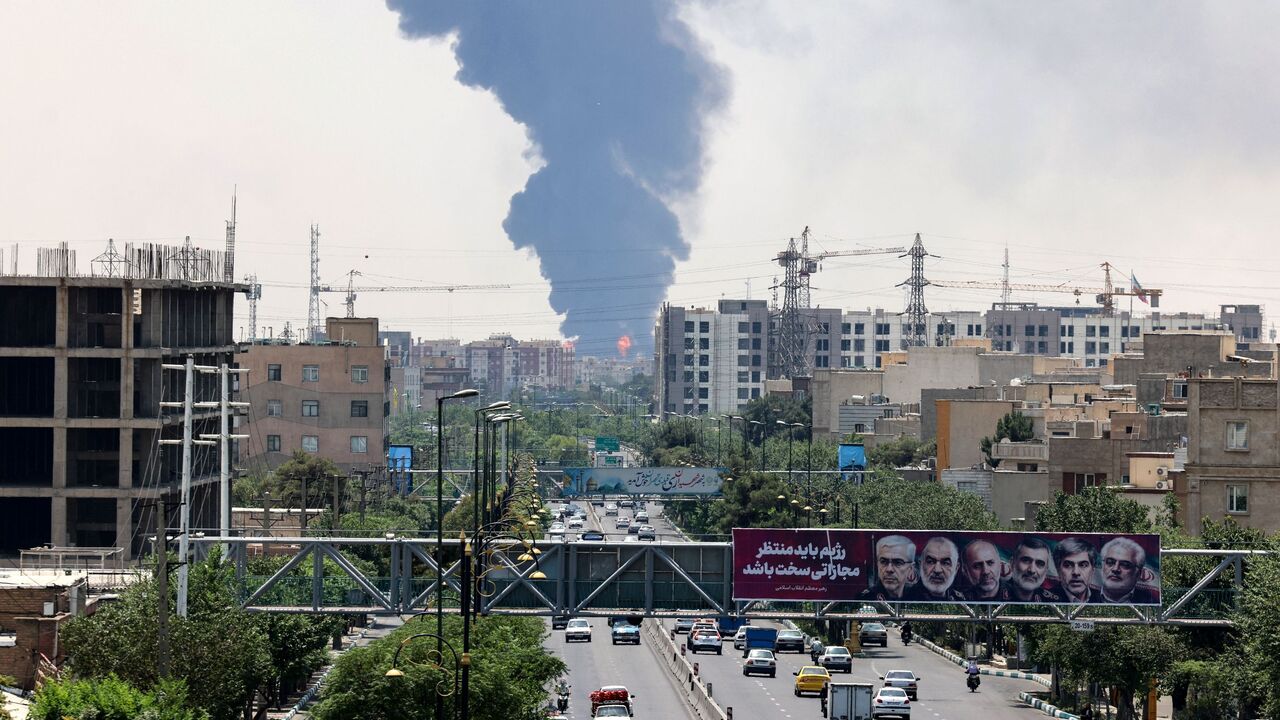 Portraits of Iranian military generals and nuclear scientists, killed in Israel's June 13 attack are displayed above a road, as a plume of heavy smoke and fire rise from an oil refinery in southern Tehran, after it was hit in an overnight Israeli strike, on June 15, 2025.