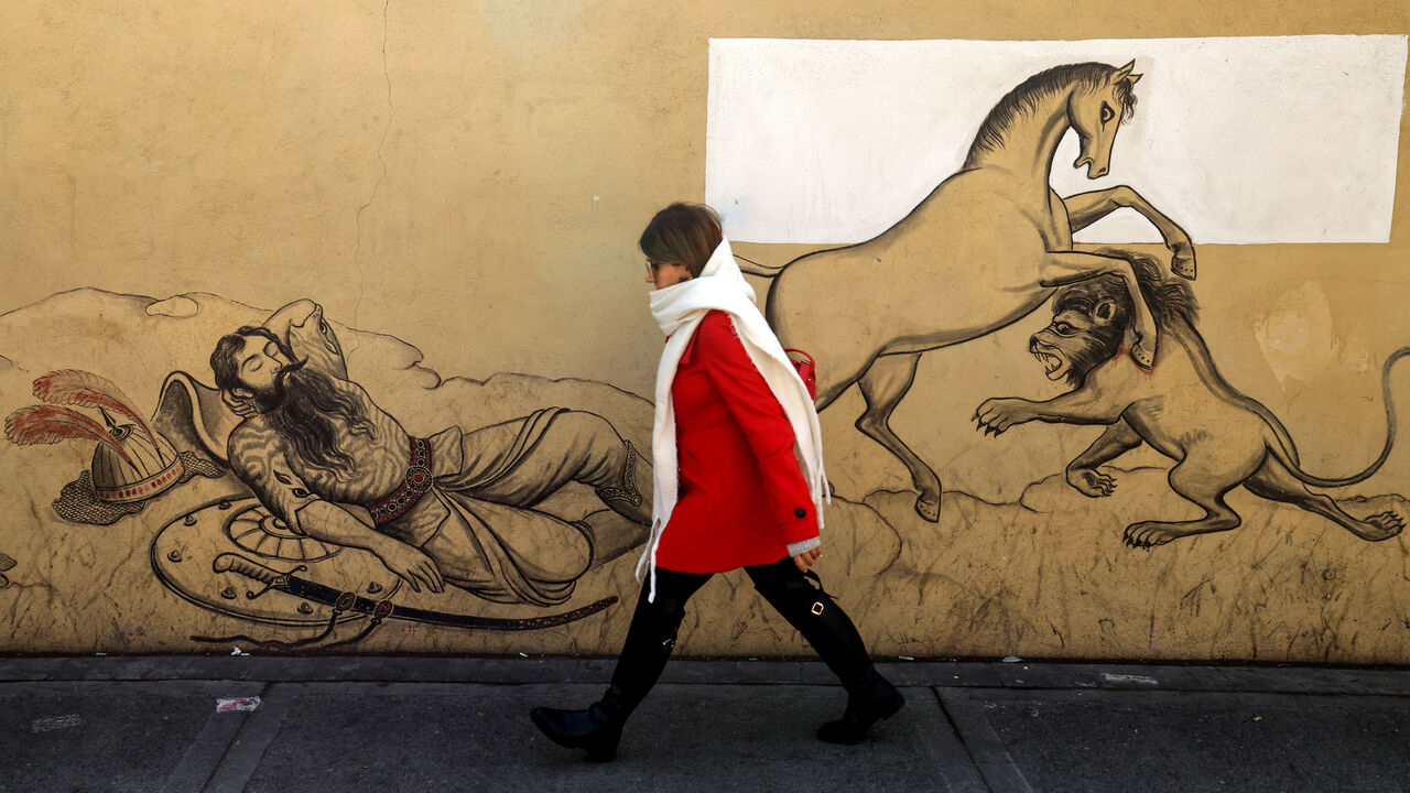 An Iranian woman walks past a mural depicting a scene from the Iranian national epic "Shahnameh" in the capital Tehran on January 6, 2026. Iran's economy has been hit hard by tough international sanctions, with the national currency, the rial, losing more than a third of its value against the US dollar over the past year and inflation in double digits. (Photo by ATTA KENARE / AFP via Getty Images)
