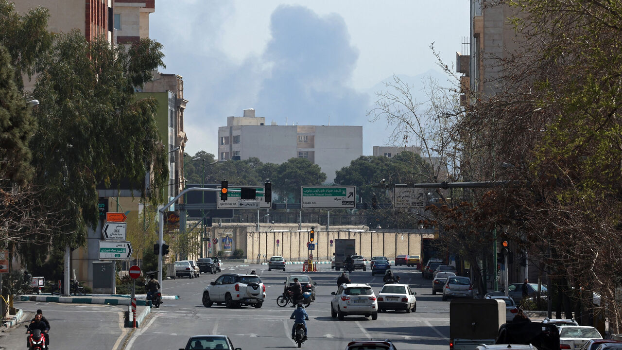 Commuters make their way along a busy street against the backdrop of a cloud of smoke rising after airstrikes in central Tehran on March 4, 2026. 