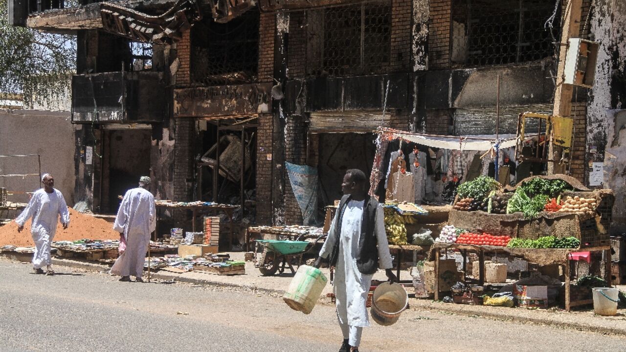 Men walk past street vendors' stands set up beneath a damaged building in the capital Khartoum on the third anniversary of the start of the war 