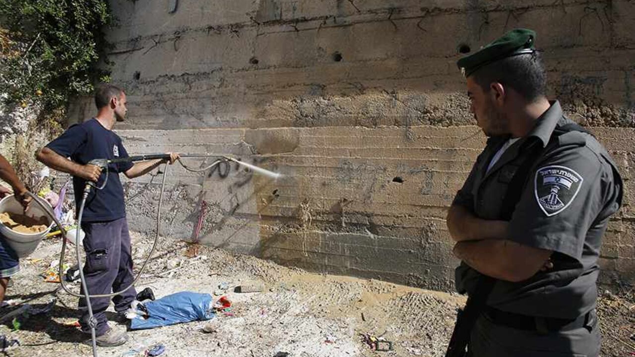 ISRAEL-PALESTINIANS/VIOLENCE A Israeli border police officer looks as a municipality worker washes off graffiti sprayed on a wall in the Arab neighborhood of Beit Hanina in East Jerusalem June 24, 2013. Graffiti was sprayed and tires were slashed on 21 cars overnight in Beit Hanina in what appeared to be another in a series of "Price Tag" attacks by suspected Jewish militants. Police opened an investigation. The graffiti read: "We will not remain silent in the face of the stone-throwing". REUTERS/Ammar Awad (JERUSALEM - Tags: POLITICS