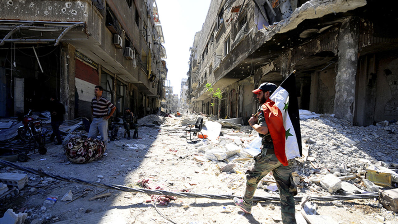 A member of forces loyal to Syria's President Bashar al-Assad holds the Syrian flag as he walks on debris of damaged buildings in al-Dukhaneya neighbourhood near Damascus A member of forces loyal to Syria's President Bashar al-Assad holds the Syrian flag as he walks on debris of damaged buildings in al-Dukhaneya neighbourhood near Damascus, after the forces regained control of the area from rebel fighters October 7, 2014. REUTERS/Omar Sanadiki (SYRIA - Tags: POLITICS CIVIL UNREST CONFLICT) - RTR499FJ