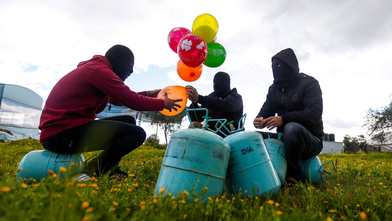 1199830046 Masked Palestinians prepare to attach balloons to a gas canister before releasing it near Gaza's Bureij refugee camp, along the Israel-Gaza border fence, on February 10, 2020. (Photo by MAHMUD HAMS / AFP) (Photo by MAHMUD HAMS/AFP via Getty Images)