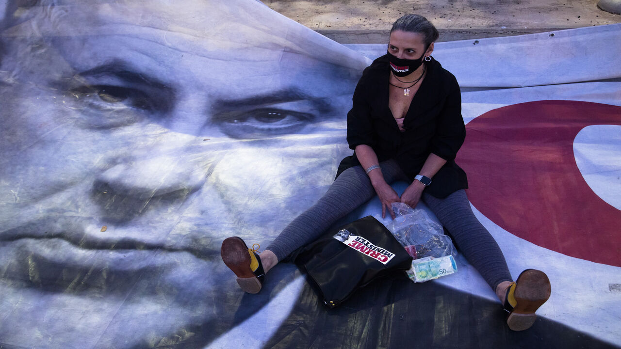 1210403955 TEL AVIV, ISRAEL - APRIL 19: A woman sits on a placard showing Israeli Prime Minister Benjamin Netanyahu, as she protests at a rally in Rabin Square on April 19, 2020 in Tel Aviv, Israel. Thousands of Israelis gather at an Anti-Corruption rally under coronavirus restrictions, decrying proposed unity government talks between Israeli Prime Minister Benjamin Netanyahu, and Blue and White Party leader Benny Gantz. (Photo by Amir Levy/Getty Images)