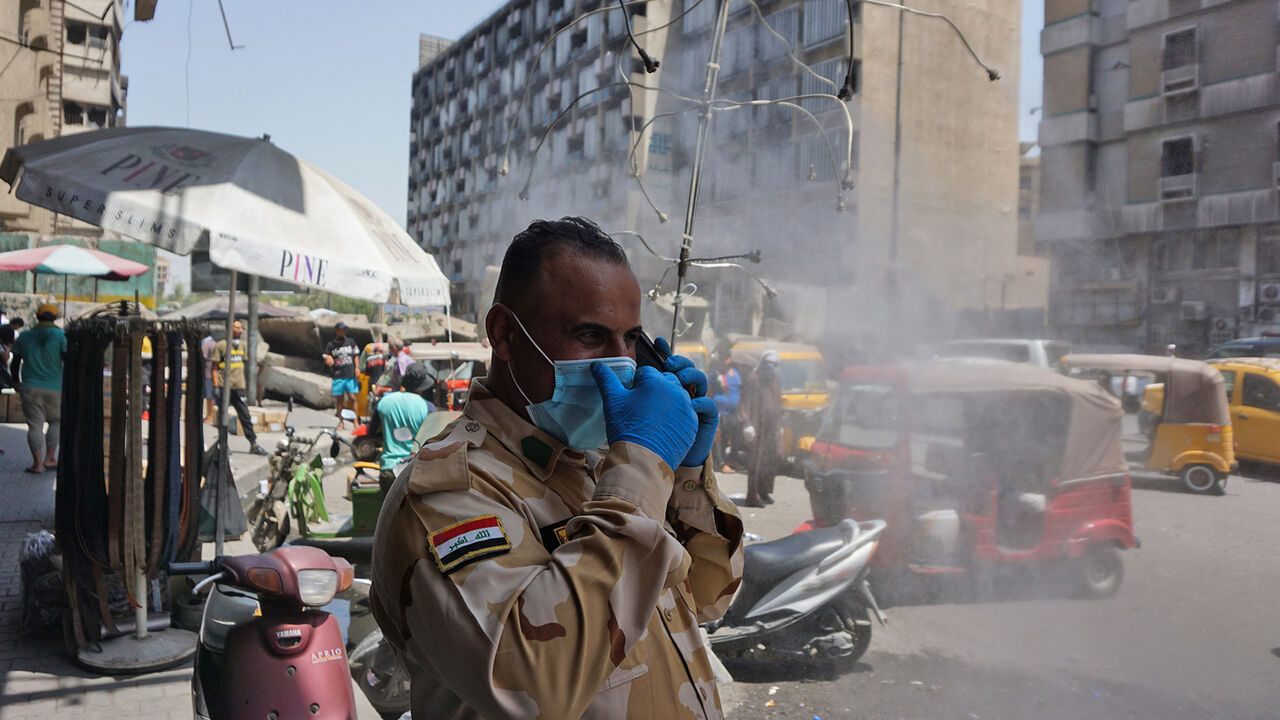 1225241922 An Iraqi policeman, wearing a protective mask and gloves due to the COVID-19 pandemic, stands by a water sprinkler on a hot summer day in Baghdad on July 7, 2020. (Photo by SABAH ARAR / AFP) (Photo by SABAH ARAR/AFP via Getty Images)
