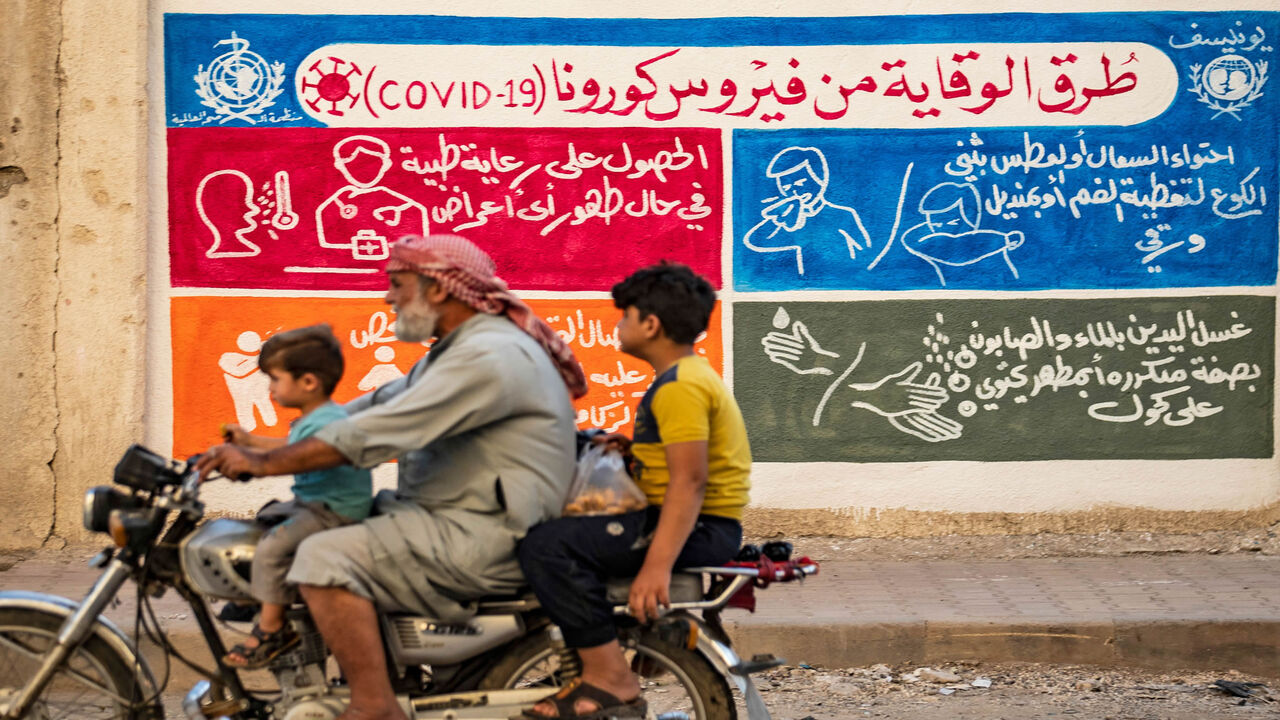 1228070822 Syrians ride a motorbike past a mural painted as part of an awareness campaign by the United Nations International Children's Emergency Fund (UNICEF) and World Health Organization (WHO) intitative, bearing instructions on protection from COVID-19 in the Kurdish-majority city of Qamishli of Syria's northeastern Hasakeh province on August 16, 2020, after a spike in infections in the area. (Photo by Delil SOULEIMAN / AFP) (Photo by DELIL SOULEIMAN/AFP via Getty Images)