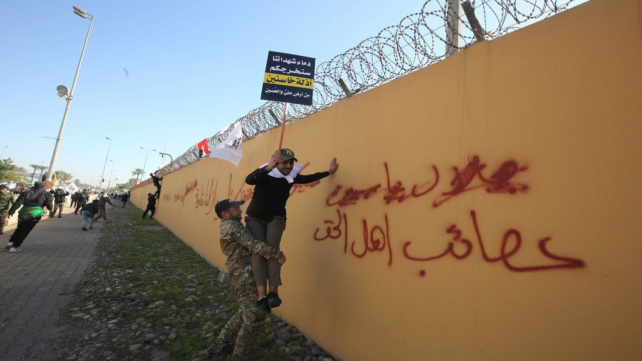 1191101216 A member of an Iraqi Shiite paramilitary group helps a fellow protesters holding a placard denouncing the United States climb on the outer wall of the US embassy in Baghdad's Green Zone during an angry demonstration on December 31, 2019 to denounce weekend US air strikes that killed Iran-backed fighters in Iraq. Arabic graffiti reads: "Assaib Ahl al-Haq" -- a pro-Iran Iraqi Shiite group whose leaders were recently hit with US sanctions. - It was the first time in years protesters have been able to reach the