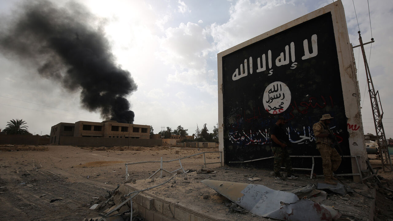 869813922 Iraqi fighters of the Hashed al-Shaabi (Popular Mobilisation units) stand next to a wall bearing the Islamic State (IS) group flag as they enter the city of al-Qaim, in Iraq's western Anbar province near the Syrian border as they fight against remnant pockets of Islamic State group jihadists on November 3, 2017. / AFP PHOTO / AHMAD AL-RUBAYE (Photo credit should read AHMAD AL-RUBAYE/AFP via Getty Images)