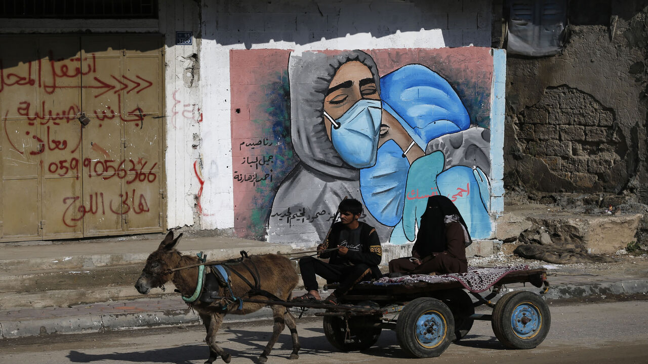 1229639648 A Palestinian man and his wife ride a donkey cart past street art showing doctors mask-clad due to the COVID-19 coronavirus pandemic in the Nusseirat refugee camp in the central Gaza Strip on November 16, 2020. (Photo by MOHAMMED ABED / AFP) (Photo by MOHAMMED ABED/AFP via Getty Images)