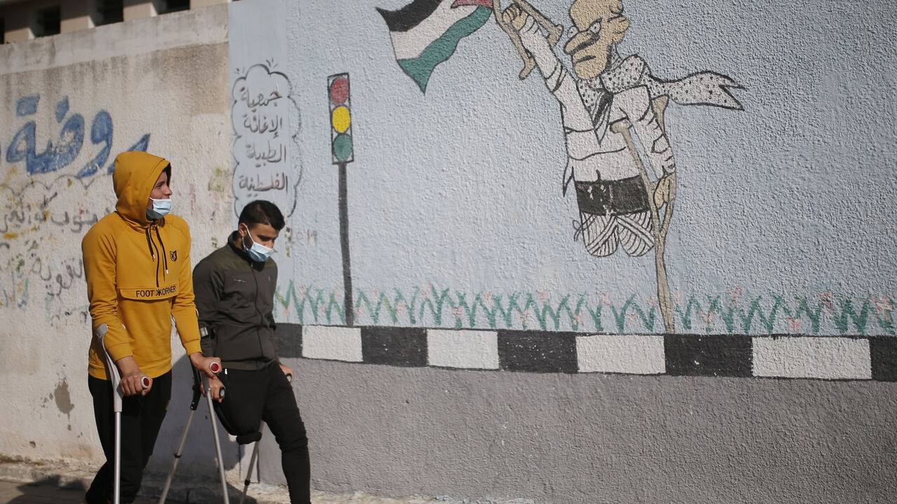 1229922261 Two Palestinian youths on crutches wearing a protective mask due to the Covid-19 pandemic, walk past graffiti depicting a disabled man carrying a national flag, during an event marking the International Day of Persons with Disabilities in Gaza City on December 3, 2020. (Photo by MOHAMMED ABED / AFP) (Photo by MOHAMMED ABED/AFP via Getty Images)