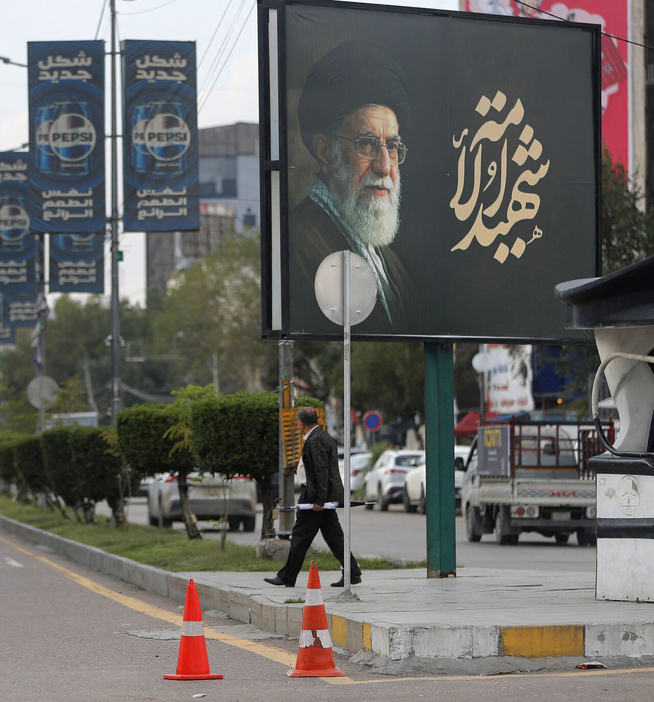 An Iraqi police officer stands guard as a man walks beneath a billboard bearing a portrait of Iran's slain supreme leader Ayatollah Ali Khamenei in Baghdad on March 25, 2026. Iraq will submit a formal complaint to the United Nations Security Council over strikes in its territory, the office of the prime minister said on March 25, following an attack in the country's west that killed seven security personnel. Iraq has been drawn into the Middle East war triggered by the US-Israel attack on its neighbour Iran