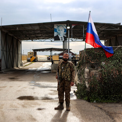 TOPSHOT - A Syrian rebel fighter (C) looks on as he stands at an inspection checkpoint for incoming vehicles before a Russian flag and Russian soldiers behind manning the entrance of the Russian-leased Syrian military base of Hmeimim in Latakia province in western Syria on December 29, 2024. (Photo by AAREF WATAD / AFP) (Photo by AAREF WATAD/AFP via Getty Images)