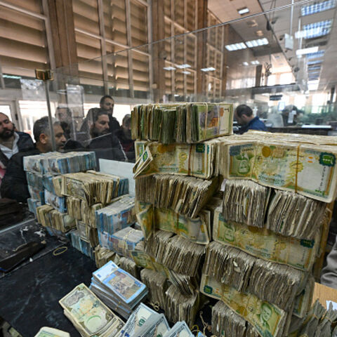  People queue in front of a teller next to a pile of banknotes of different currencies at a commercial bank affiliated with Syria's central bank in Damascus on December 16, 2024. Governments worldwide are stepping up efforts to engage with Syria's new interim rulers, just over a week after Islamist-led rebels ousted president Bashar al-Assad, ending decades of brutal rule and civil war. (Photo by Louai Beshara / AFP) / "The erroneous CAPTION appearing in the metadata of this photo by Louai Beshara has been 