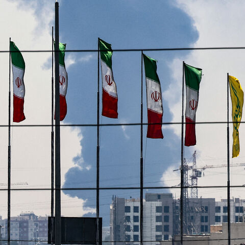 Flags flutter along a bridge as a plume of heavy smoke and fire rise from an oil refinery in southern Tehran, after it was hit in an overnight Israeli strike, on June 15, 2025. Iran fired a fresh barrage of missiles at Israel, state television announced early on June 15, as the rivals exchanged fire for a third day. (Photo by ATTA KENARE / AFP) (Photo by ATTA KENARE/AFP via Getty Images)