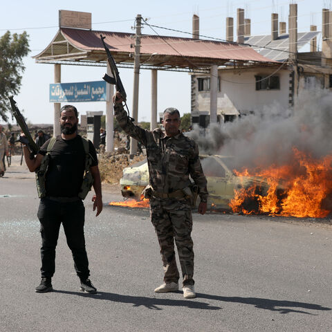 Two armed men gesture as a car burns near the site of clashes between Druze and Bedouin factions on July 14, 2025 in Suwayda, Syria.