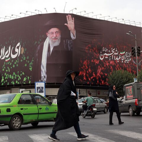 People cross a street next to a billboard bearing the portrait of Iranian Supreme Leader Ayatollah Ali Khamenei and a quote that reads "Sing Oh Iran" in Tehran on July, 9, 2025. 