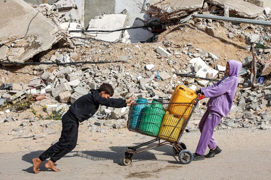 A girl pulls while a boy pushes a shopping-cart loaded with filled-up water containers past a mound of rubble and debris in Gaza City