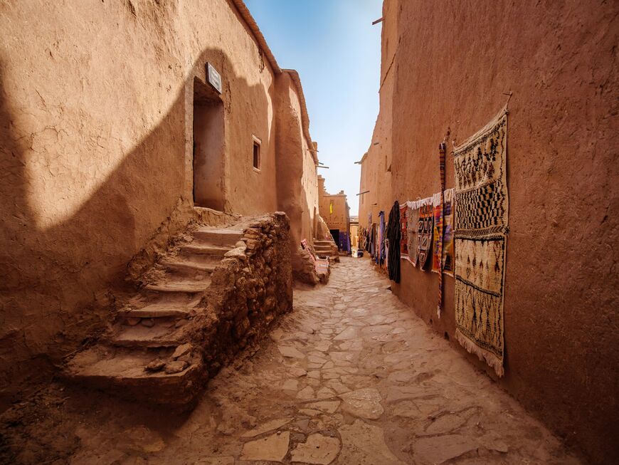 Empty street in the Ait Benhaddou ksar (Aït Benhaddou Kasbah), a famous berber fortified village, with rugs and clothes of a souvenir shop of a street vendor hanged on an earthen clay wall, UNESCO World Heritage Site - Ouarzazate - Morocco. - stock photo